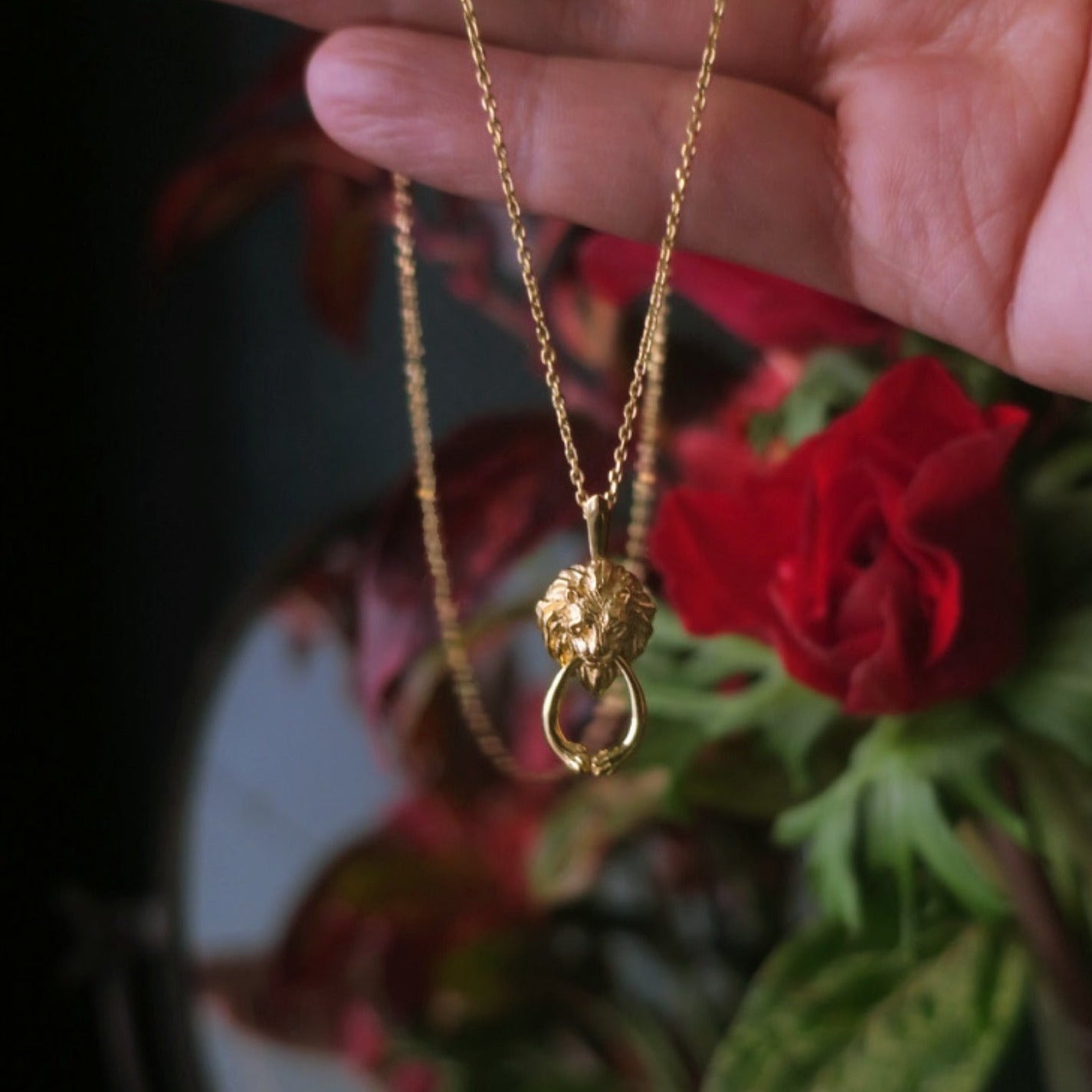 Gold lion door-knocker necklace styled near a vintage mirror and anemone flowers, highlighting the antique-inspired design and hand-sculpted lion pendant.