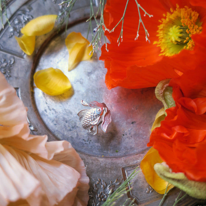 Sterling silver goldfish ring displayed among vibrant flowers – handcrafted animal jewelry from Paris, poetic composition highlighting nature and artistry.
