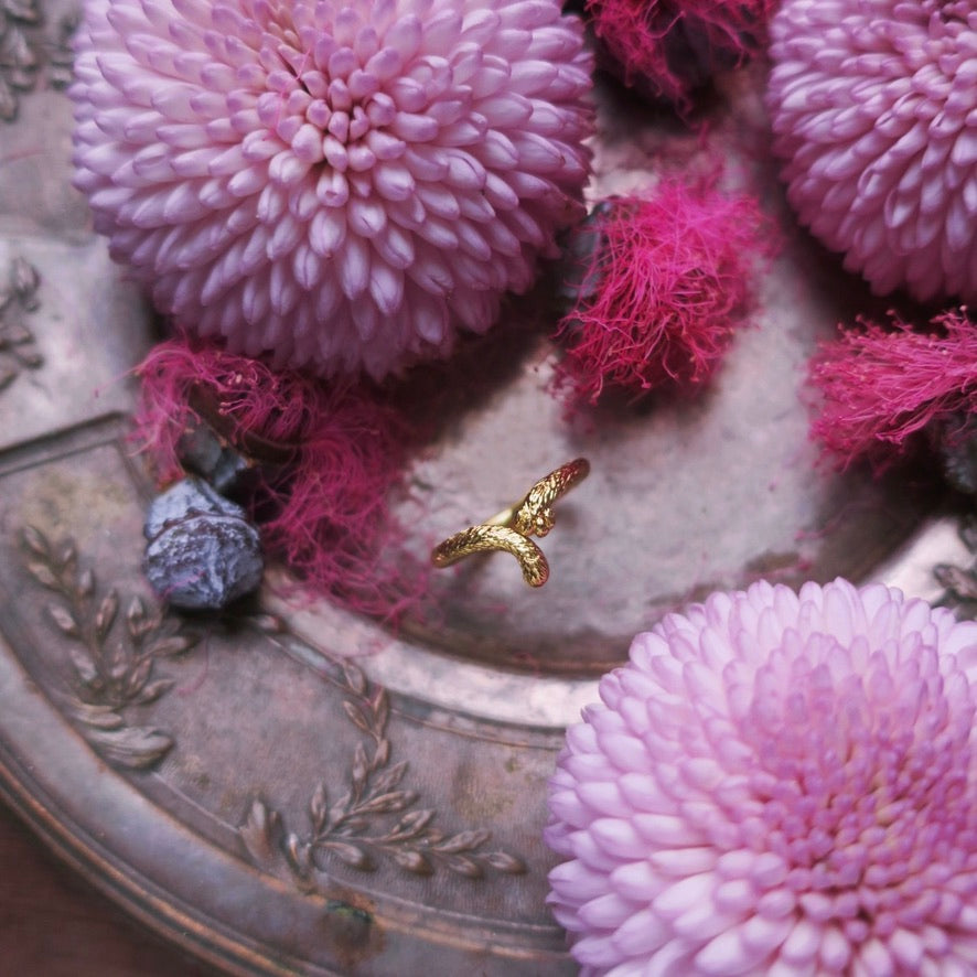 Gold cat tail ring photographed among pink florals, highlighting its delicate curved silhouette and the hidden paw pad motif. Hand-sculpted animal jewelry in Paris with fine artisanal detail.