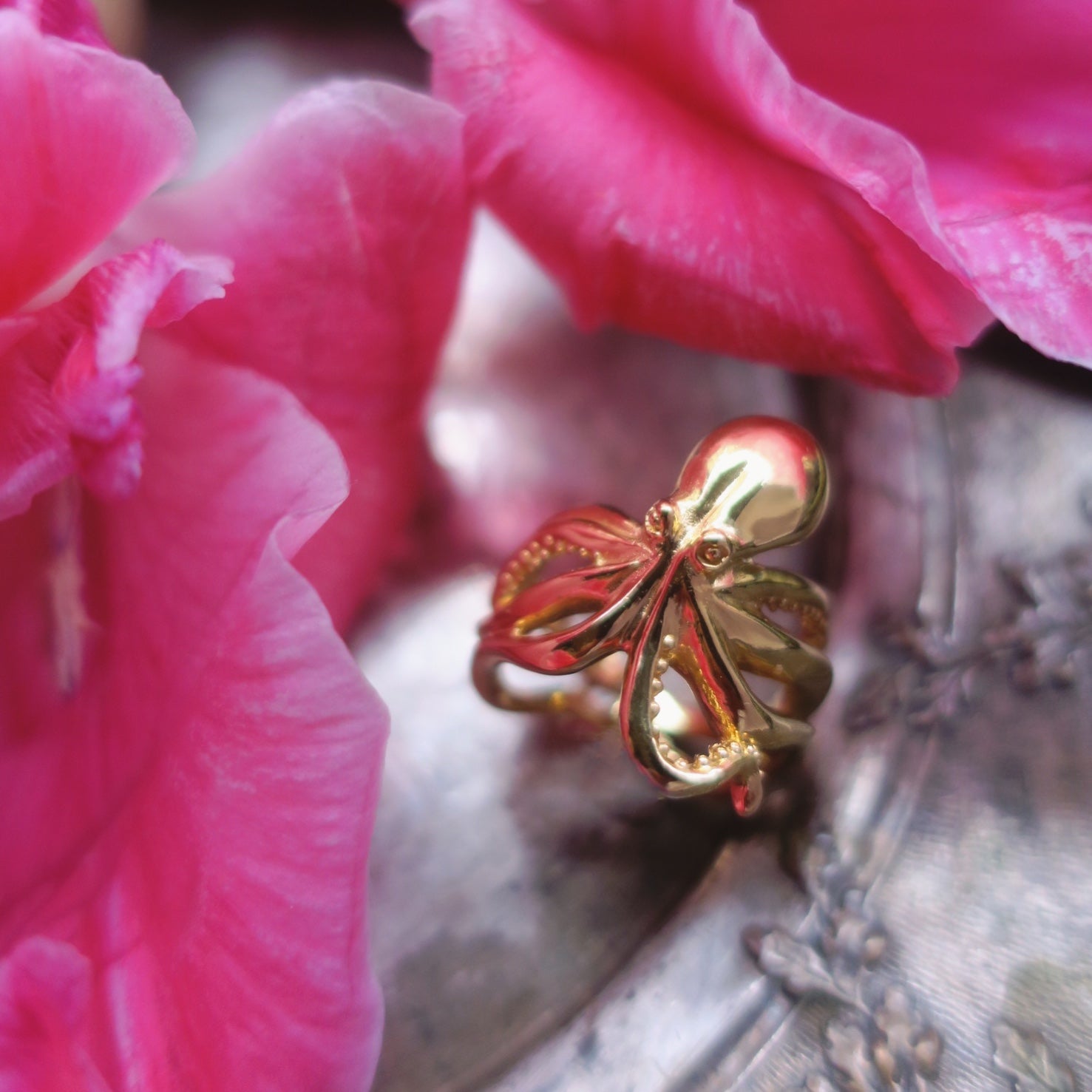 Gold octopus ring photographed with soft floral background, highlighting the fluid tentacle silhouette; ocean-themed artisanal jewelry with bold marine-inspired design.