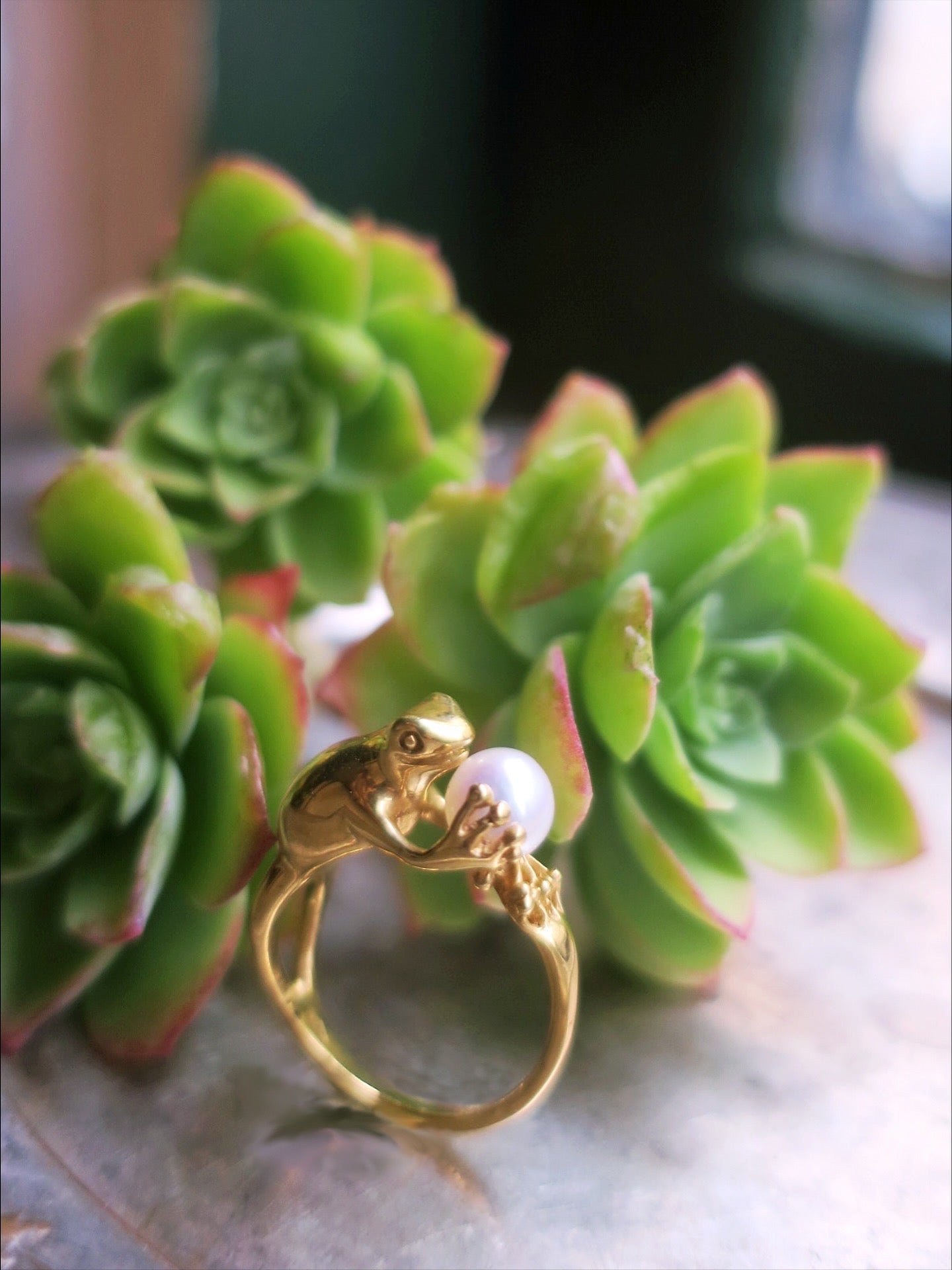 Gold-plated silver frog ring holding a freshwater pearl, photographed among green succulents — Art Nouveau-inspired handmade animal jewelry from Paris.