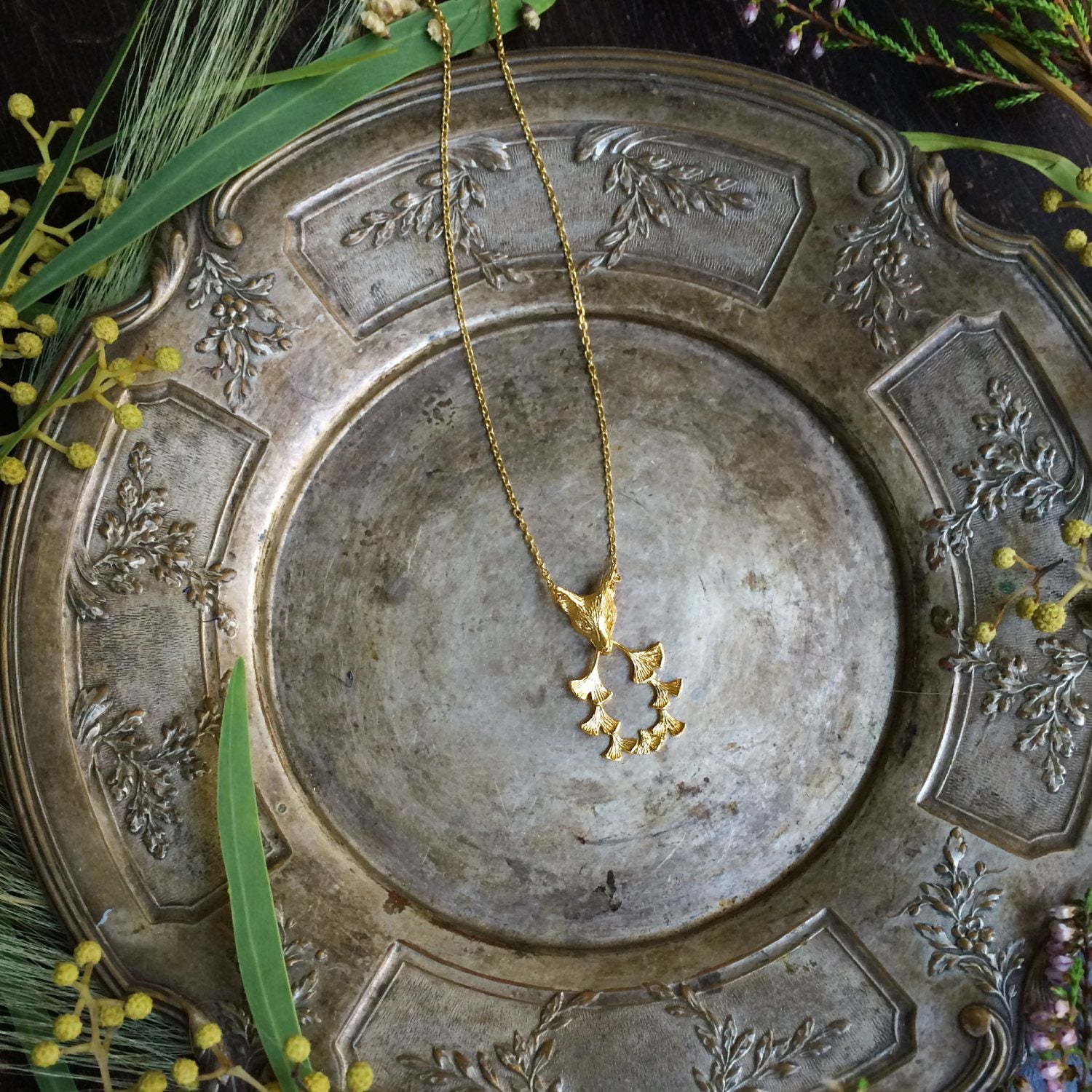 Styled Fox & Ginkgos gold plated necklace draped over vintage silver plate with greenery — warm autumn-tone product setting