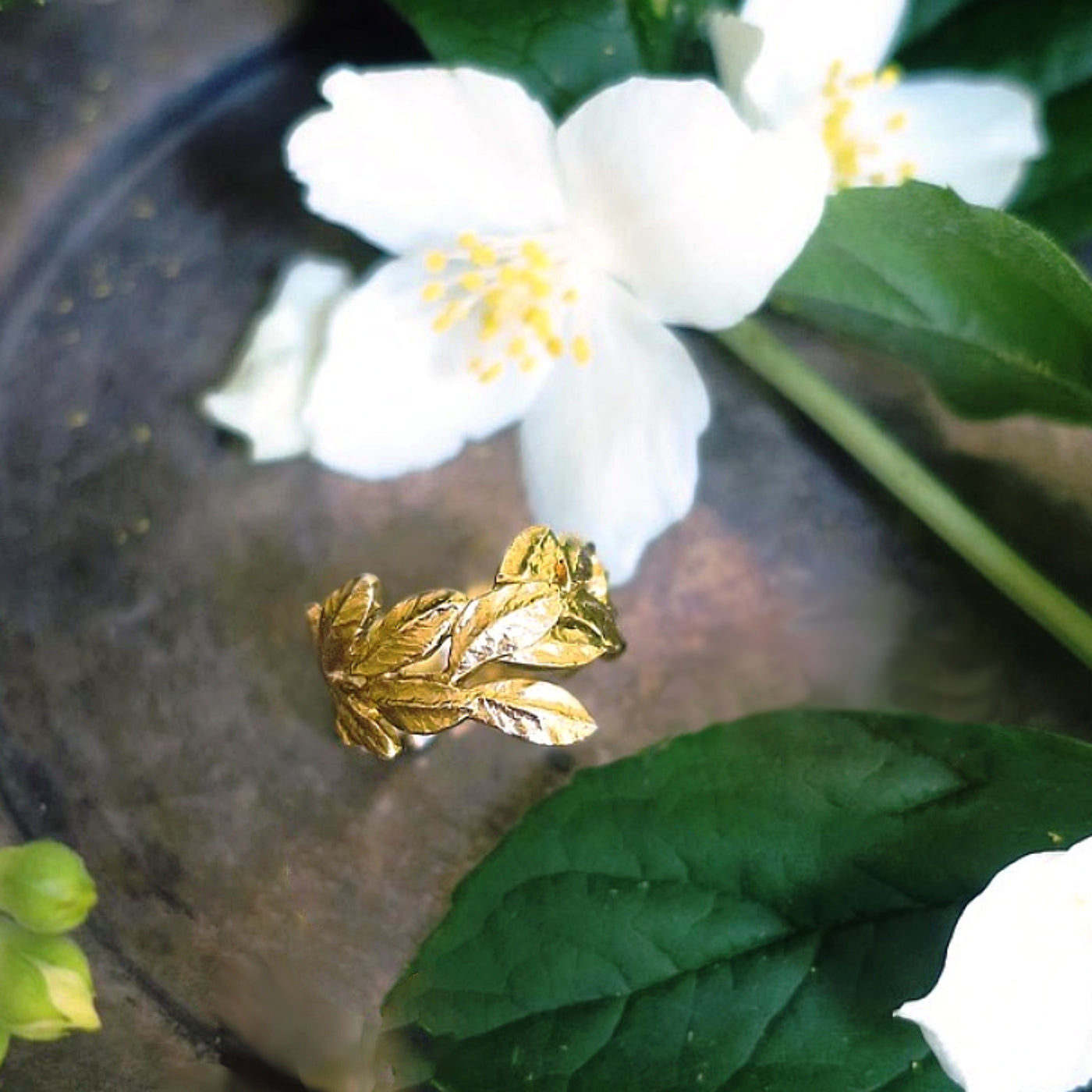 Sculptural laurel leaves ring in 18K gold-plated silver displayed beside white flowers, emphasizing hand-crafted botanical artistry and refined Parisian jewellery design.