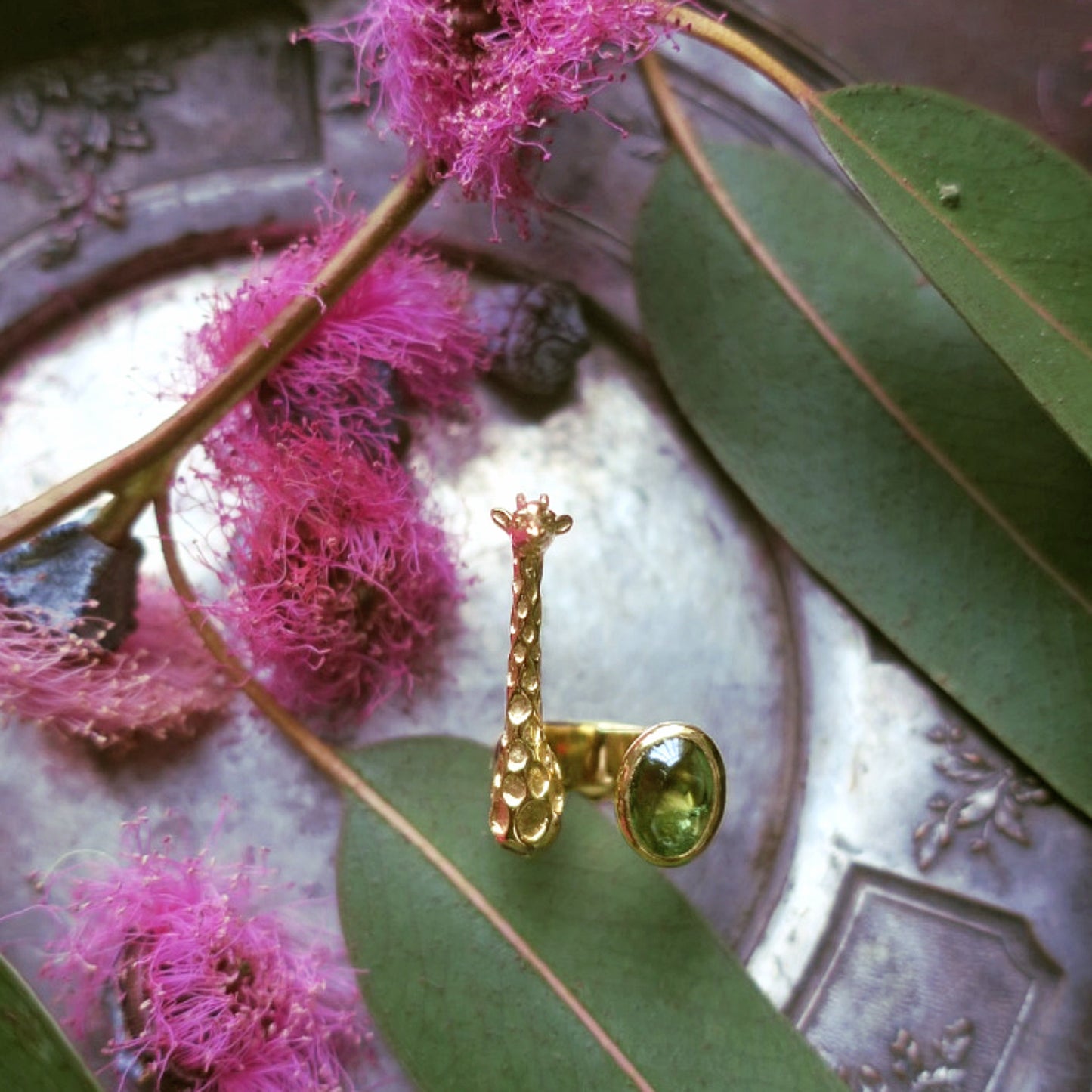 Gold giraffe ring with green tourmaline photographed on eucalyptus leaves and pink flowers, showing the natural gemstone’s inclusions
