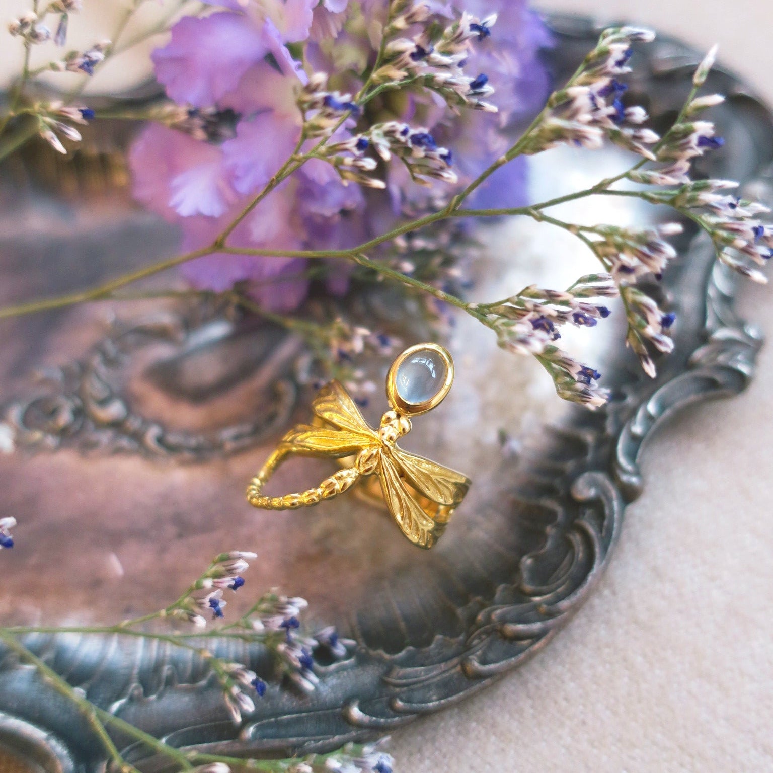 Milky aquamarine dragonfly ring placed on vintage silver tray with lilac flowers, evoking Art Nouveau romanticism and poetic craftsmanship.