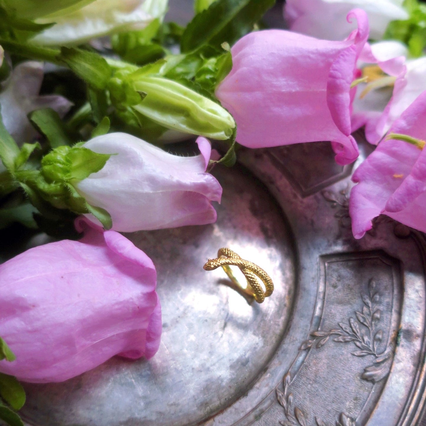 “Seeker” gold snake ring displayed among soft pink flowers, highlighting its fine craftsmanship and shimmering 18K gold finish.