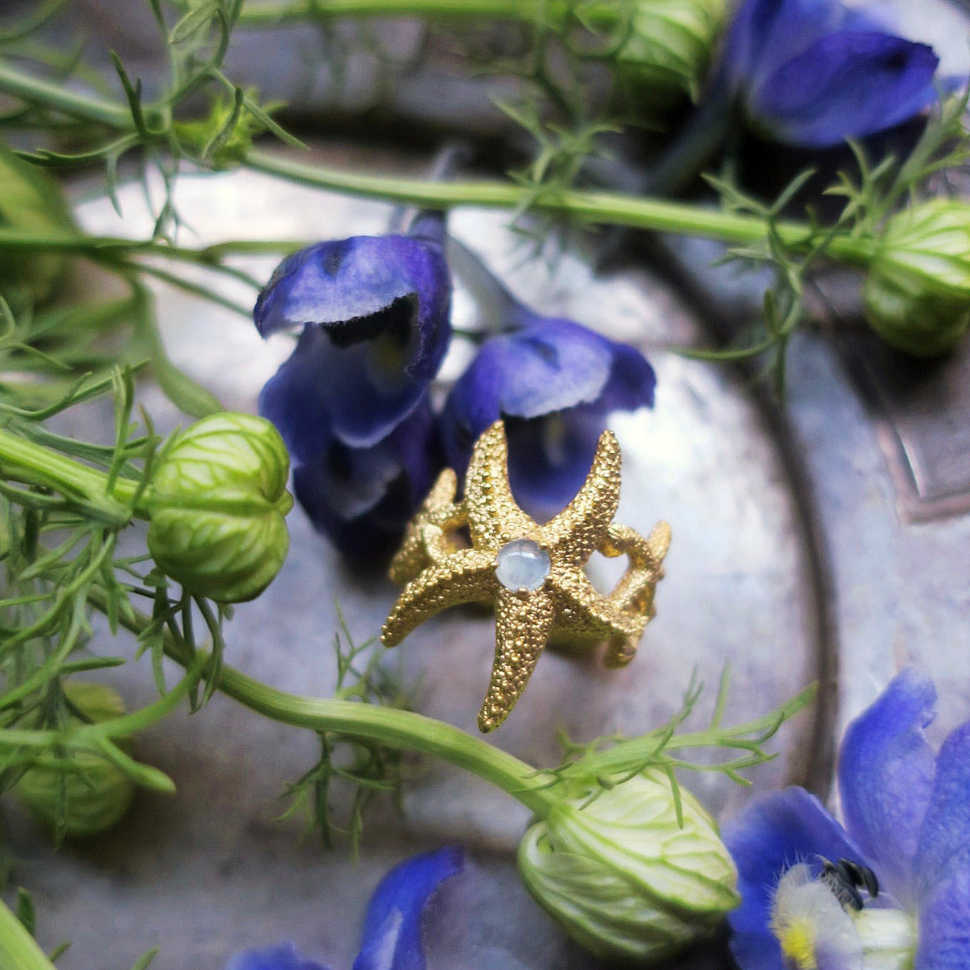 Gold starfish aquamarine ring placed among blue florals, highlighting its hand-sculpted marine texture. 18K gold-plated sterling silver with natural milky aquamarine, photographed in Paris. Ocean-inspired statement jewelry.