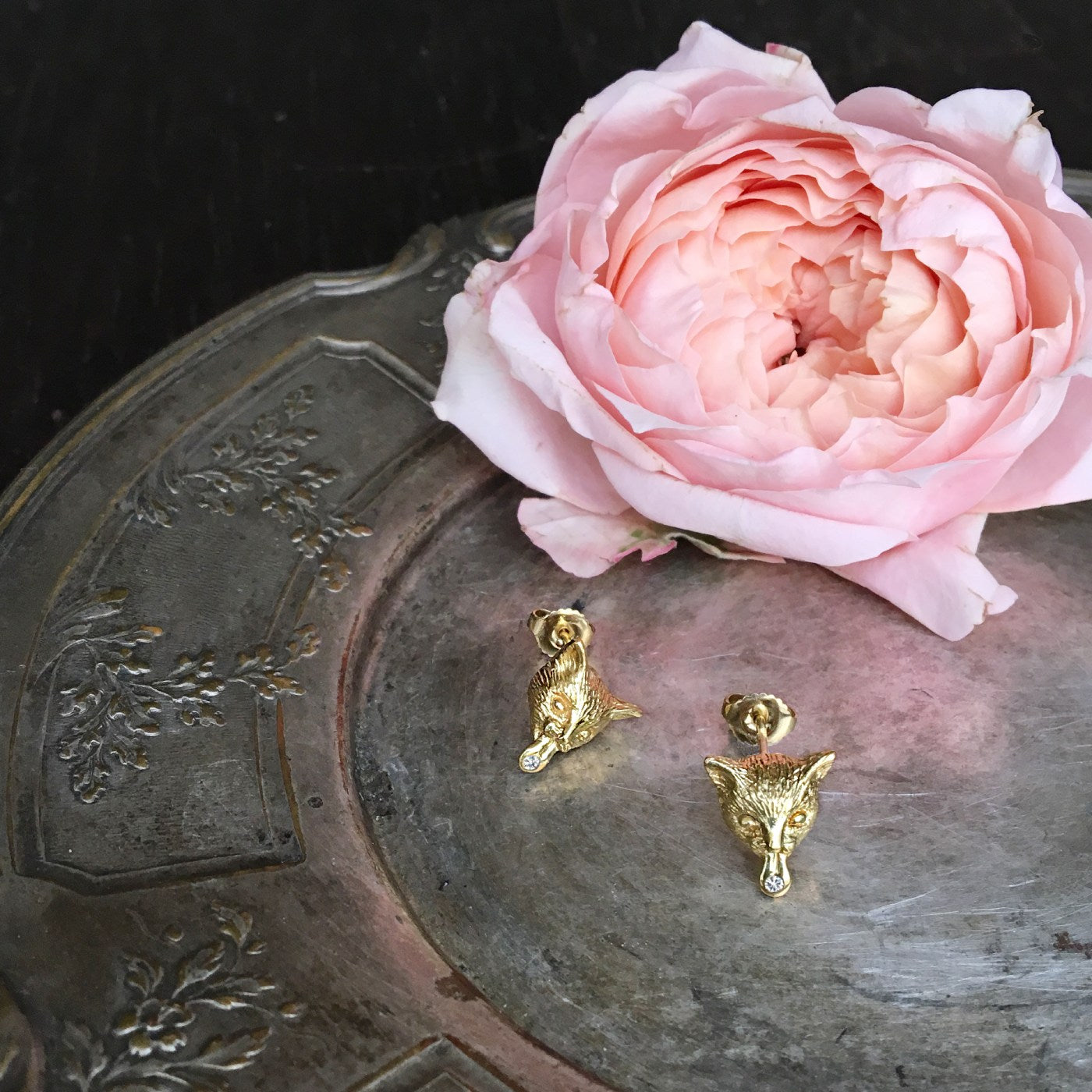 Gold-plated cat earrings with expressive faces and white sapphire tongues displayed on an antique metal tray beside a soft pink rose. Paris-made artisan animal jewelry.