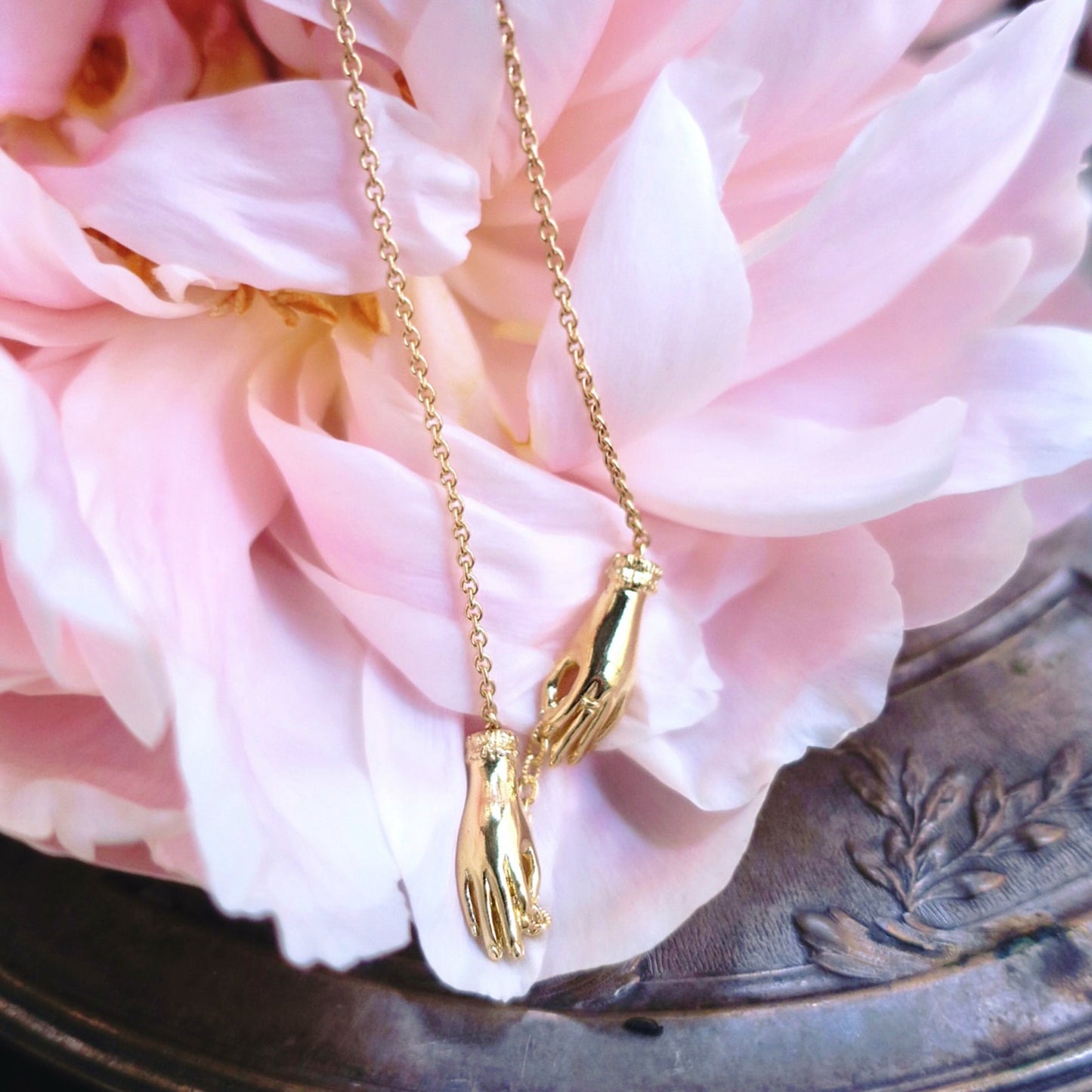 Gold sculptural hand necklace draped over a pink peony on an antique silver dish