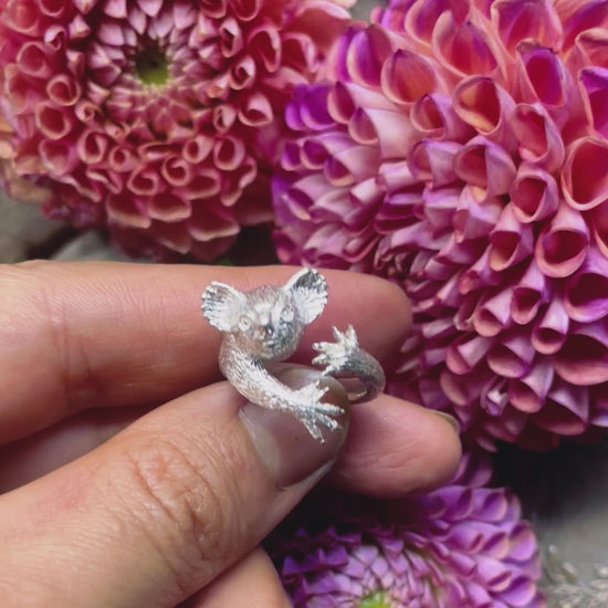 Video of a hand-sculpted sterling silver koala ring displayed over blooming dahlia flowers, showing its full 3D form, realistic details, and hugging arms — handmade animal jewelry by Fusako Koike Paris.