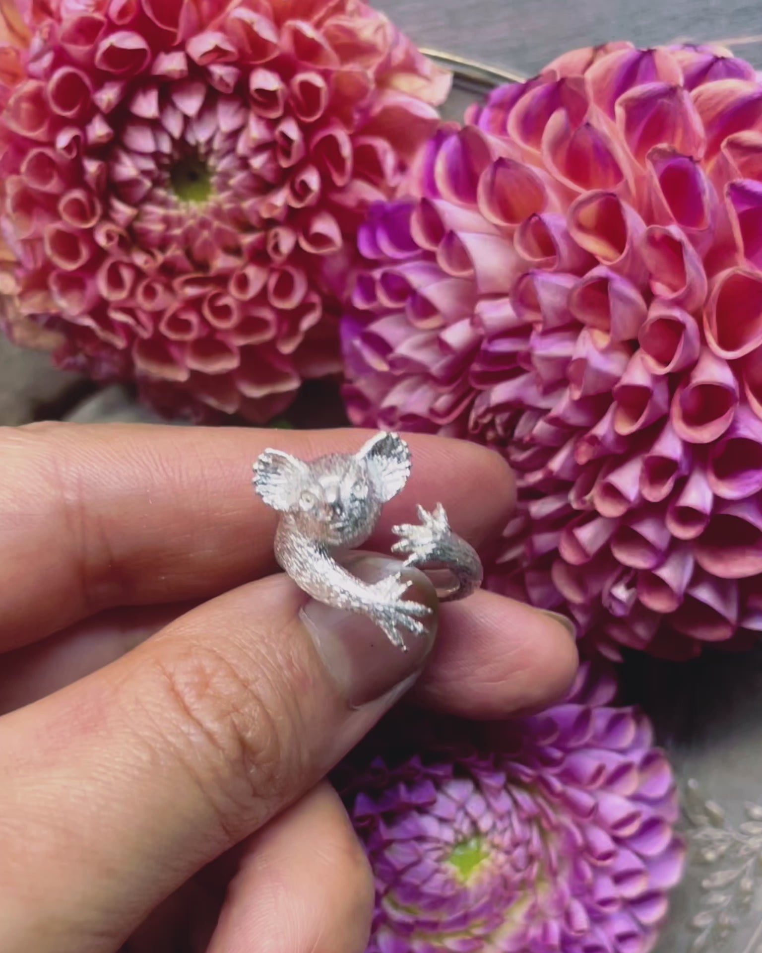 Video of a hand-sculpted sterling silver koala ring displayed over blooming dahlia flowers, showing its full 3D form, realistic details, and hugging arms — handmade animal jewelry by Fusako Koike Paris.