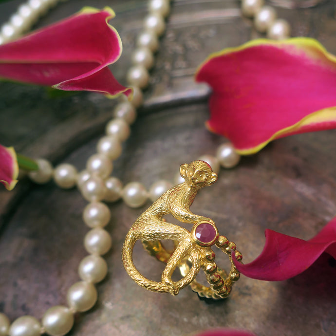 Gold-plated silver monkey ring with a rough-cut ruby, placed among pearl beads and pink lily petals — symbolic of playful luxury and antique charm.
